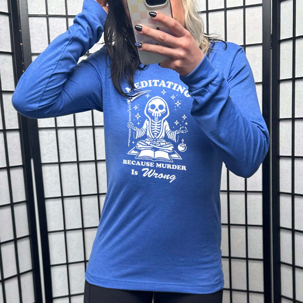 heather true royal long sleeve shirt with the text "Meditating Because Murder is Wrong" on it, with the graphic of a skeleton wearing a robe, holding a scythe, and meditating in front of an open book, with a little candle beside, and little stars scattered around