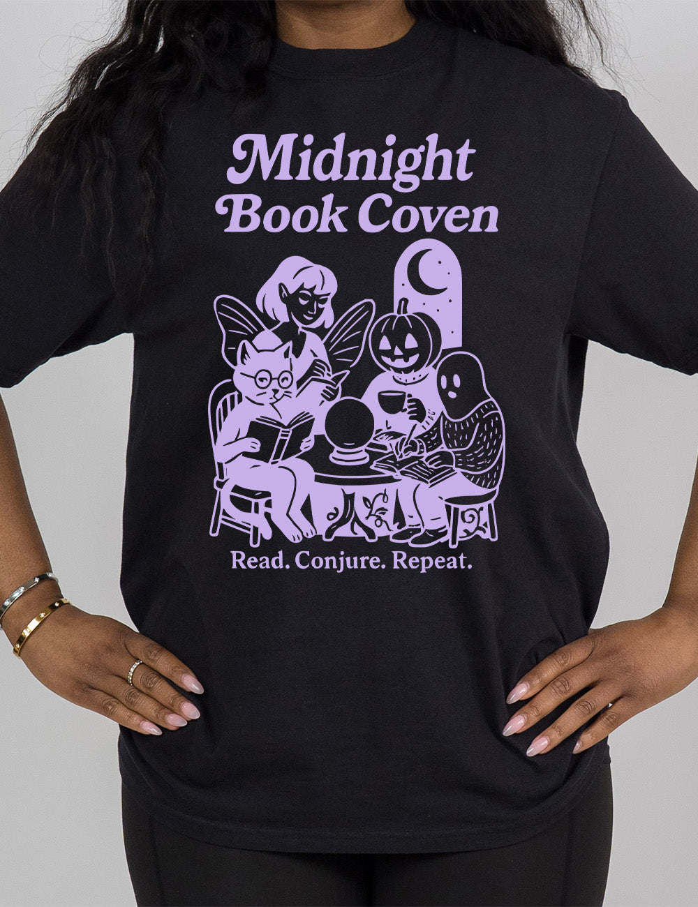 black modern tee with words "midnight book coven" above the graphic of a tombstone with a moon on it, a fairy reading a book, a pumpkin head person drinking coffee with a book, a ghost wearing a sweater and writing in a journal, and a humanoid cat wearing glasses, giving side-eye, and reading a book, all around a table with a crystal ball on top, and the words underneath saying "read. conjure. repeat."