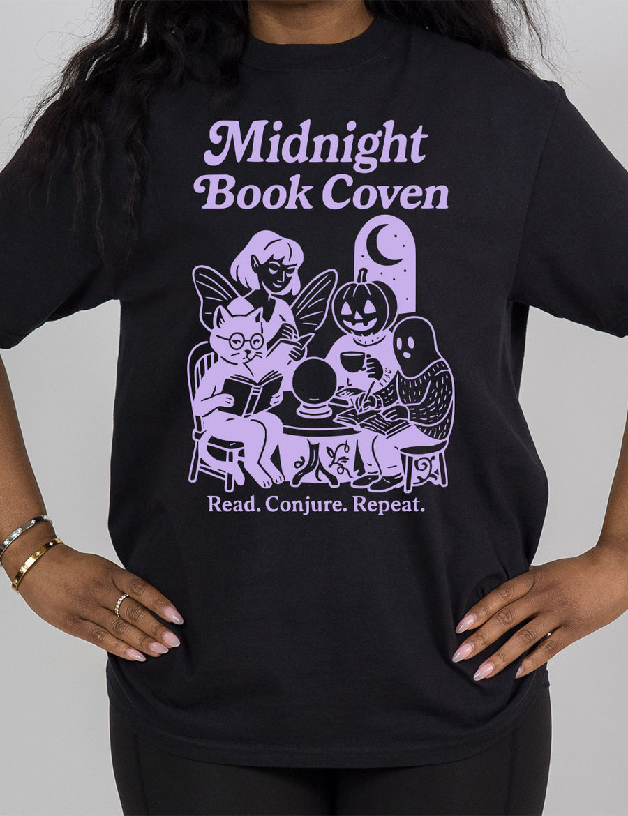 black modern tee with words "midnight book coven" above the graphic of a tombstone with a moon on it, a fairy reading a book, a pumpkin head person drinking coffee with a book, a ghost wearing a sweater and writing in a journal, and a humanoid cat wearing glasses, giving side-eye, and reading a book, all around a table with a crystal ball on top, and the words underneath saying "read. conjure. repeat."