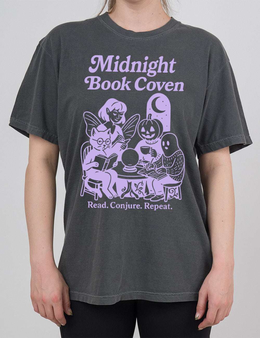 pepper modern tee with words "midnight book coven" above the graphic of a tombstone with a moon on it, a fairy reading a book, a pumpkin head person drinking coffee with a book, a ghost wearing a sweater and writing in a journal, and a humanoid cat wearing glasses, giving side-eye, and reading a book, all around a table with a crystal ball on top, and the words underneath saying "read. conjure. repeat."