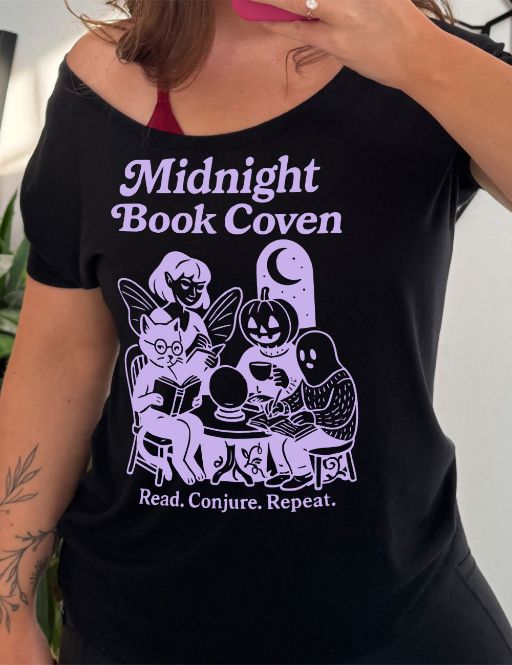 black slouchy tee with words "midnight book coven" above the graphic of a tombstone with a moon on it, a fairy reading a book, a pumpkin head person drinking coffee with a book, a ghost wearing a sweater and writing in a journal, and a humanoid cat wearing glasses, giving side-eye, and reading a book, all around a table with a crystal ball on top, and the words underneath saying "read. conjure. repeat."