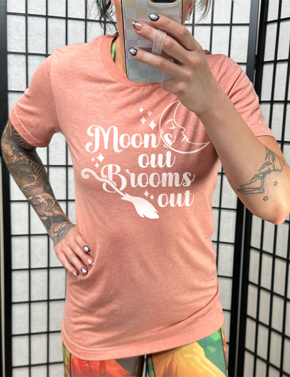 sunset unisex tee with the words "moon's out brooms out" on it, with the line graphic of a crescent moon, with a sleeping face, and the graphic of a broom, and a few scattered stars