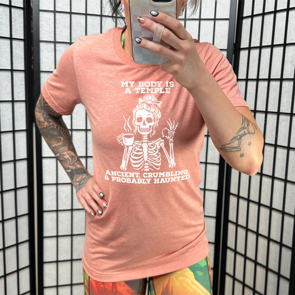 sunset unisex tee with the words "My Body Is a Temple Ancient, Crumbling, & Probably Haunted" on it, with the graphic of a skeleton with its hair in a bow, with one hand holding steaming coffee, and the other giving the OK symbol