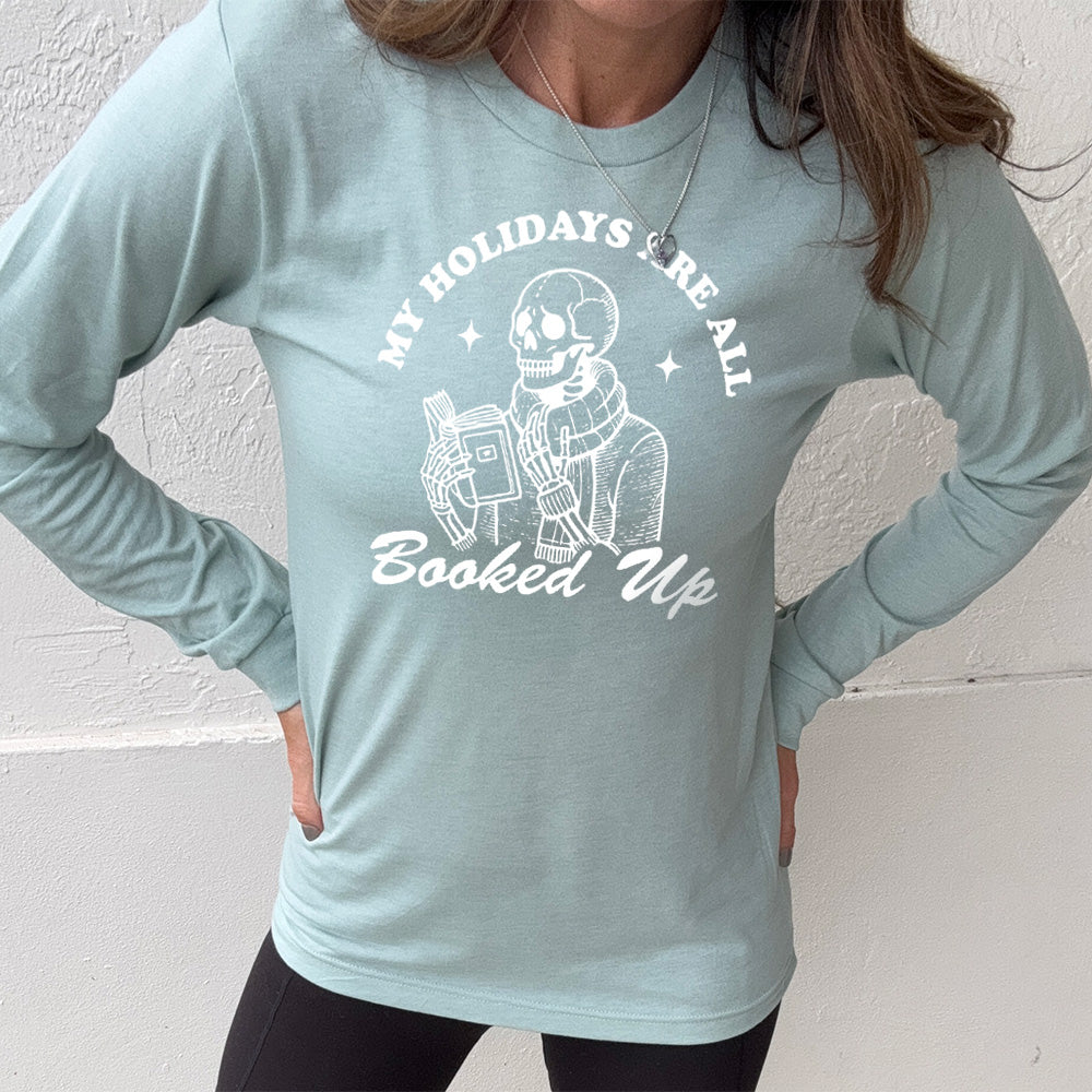 heather dusty blue long sleeve with the words "my holidays are all booked up", with the image of a skeleton dressed warmly and reading a book