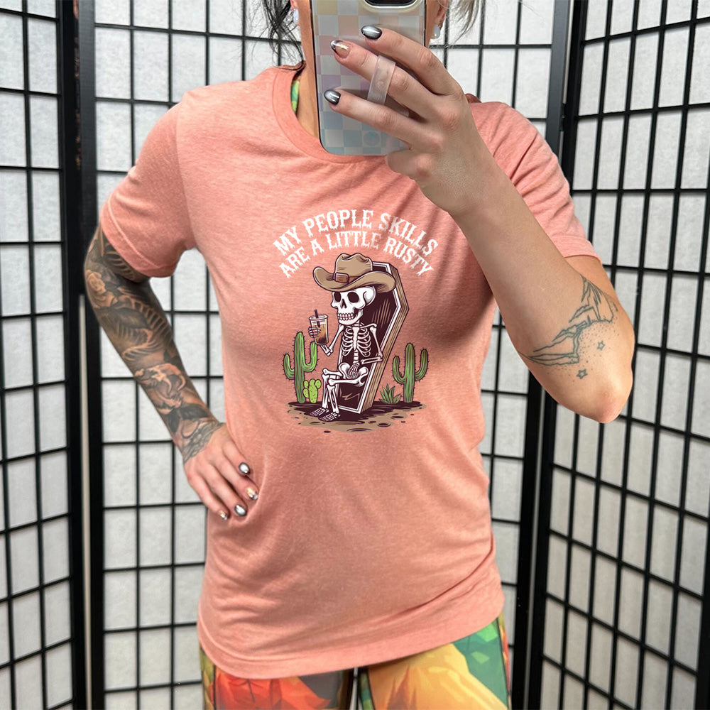 Sunset colored shirt with a skeleton sitting in a coffin graphic on it that says "My People Skills Are A Little Rusty" 