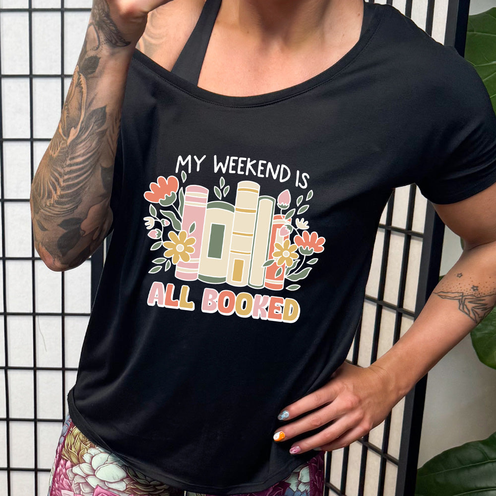 A person wearing a black slouchy tee with the text 'MY WEEKEND IS ALL BOOKED' on the front, decorated with images of books and flowers.