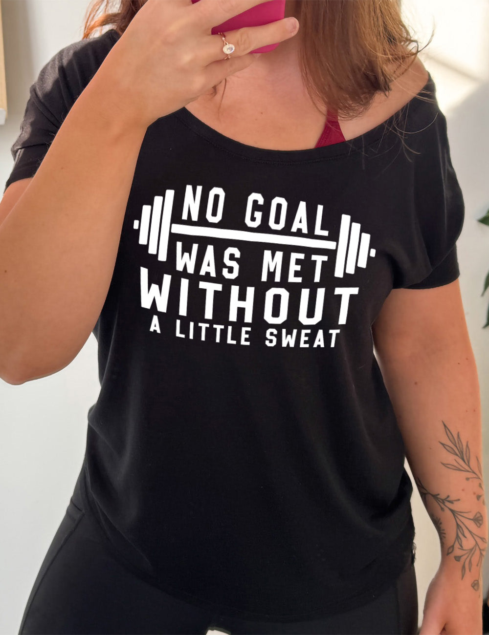 black slouchy tee with the words "NO GOAL WAS MET WITHOUT A LITTLE SWEAT" on it, with a barbell graphic after the words "no goal"