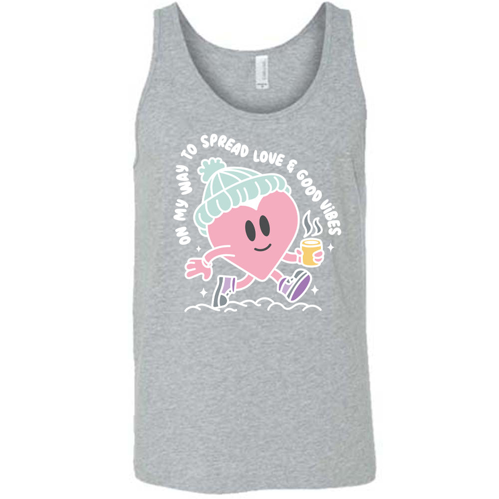 grey unisex tank with the words "on my way to spread love & good vibes" on it, with graphics of a cheerful, cartoony heart strolling along with a mug of a hot drink steaming in its hand, a hat on its head, and shoes on