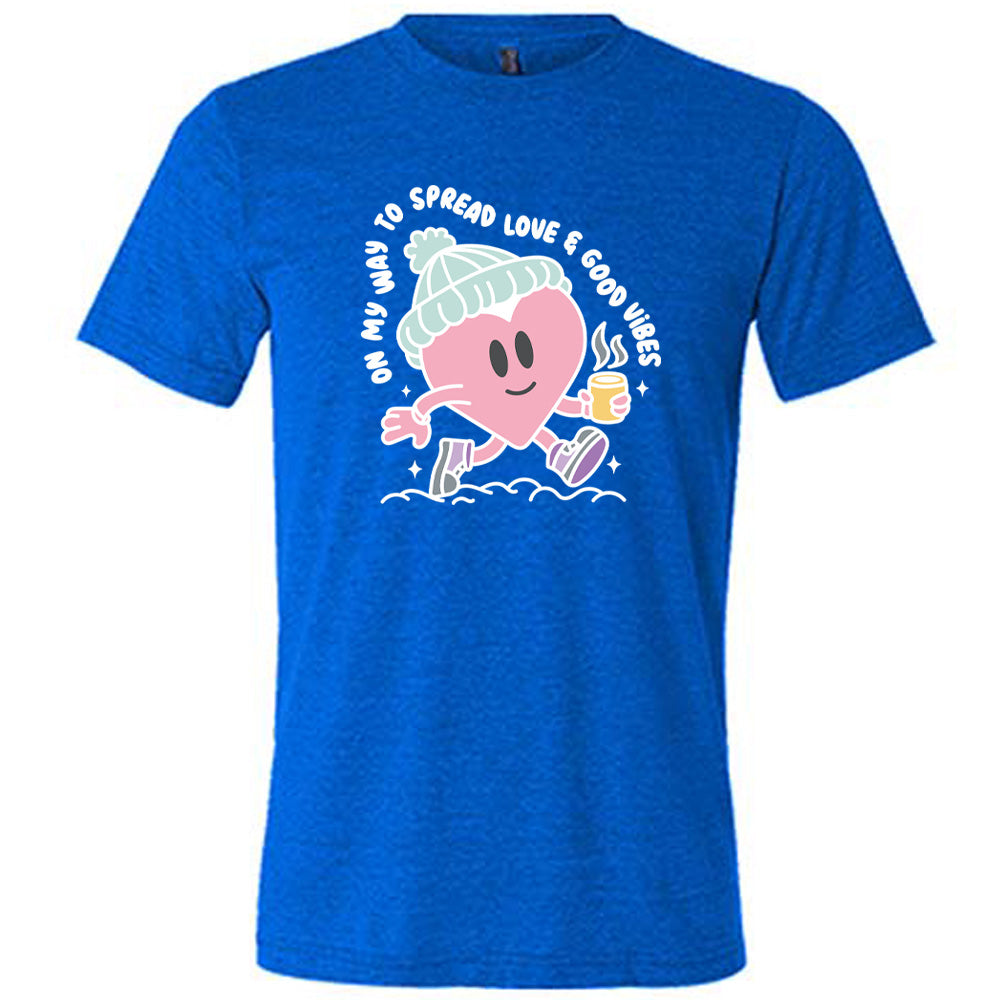 blue unisex tee with the words "on my way to spread love & good vibes" on it, with graphics of a cheerful, cartoony heart strolling along with a mug of a hot drink steaming in its hand, a hat on its head, and shoes on