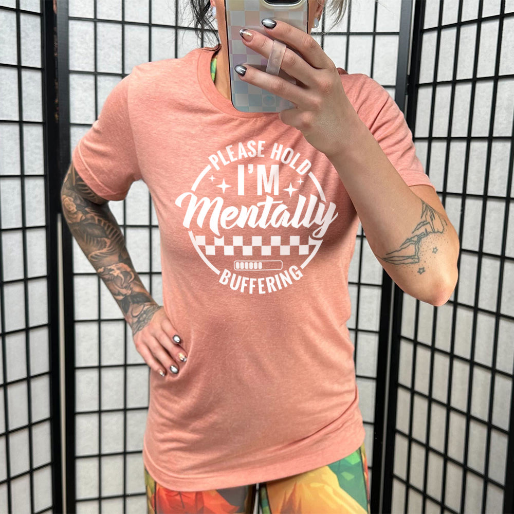 sunset unisex tee with the words "please hold I'm mentally buffering" written on it in a circle graphic, with some of the words part of the arches, and within the circle there are two layers of checkerboard as well as a buffering bar, which is about 6 out of 10 full