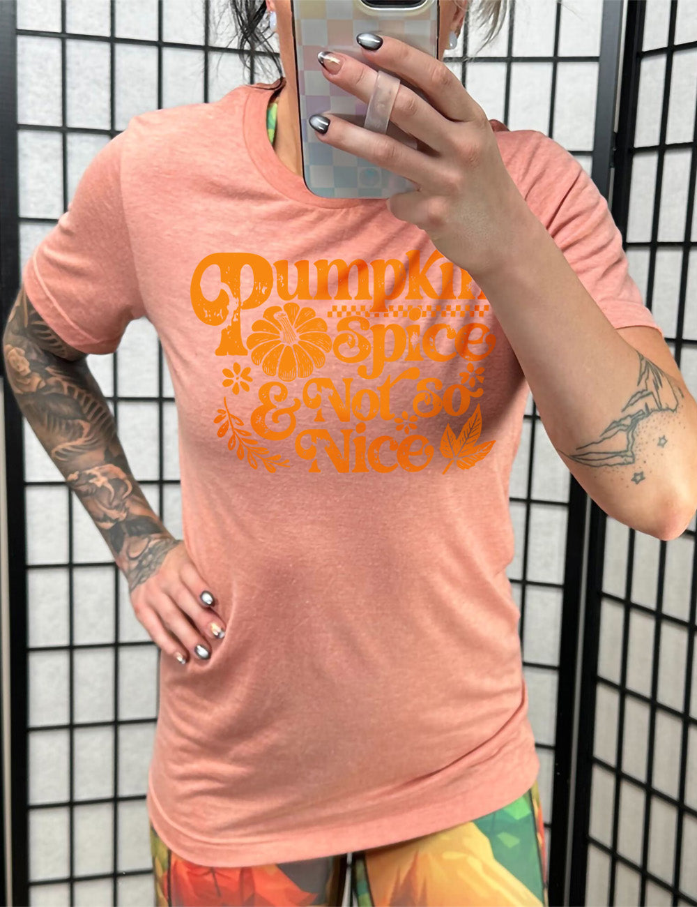 sunset unisex tee with the words "pumpkin spice & not so nice" and a top-down view of a pumpkin graphic to the left of the word "spice" and a checkered banner above the same word, and a few leaves and plants around the other words