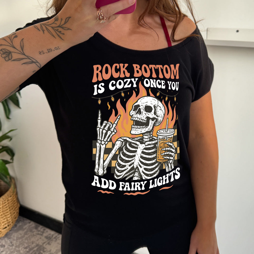 black slouchy tee with the words "rock bottom is cozy once you add fairy lights" with the graphic of a skeleton giving the rocker sign with one hand while holding an iced drink in the other, with a background of some checkerboard, some flames, and a string of fairy lights above the flames