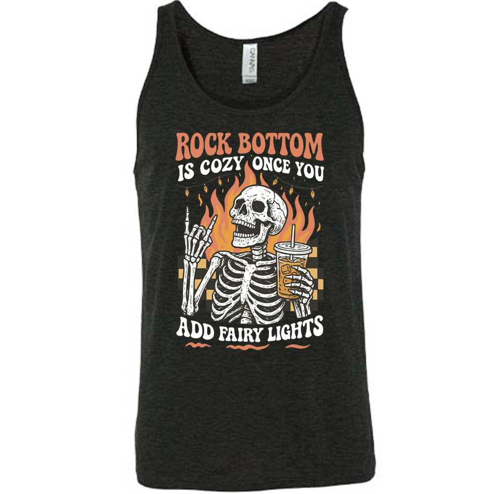 black unisex tank with the words "rock bottom is cozy once you add fairy lights" with the graphic of a skeleton giving the rocker sign with one hand while holding an iced drink in the other, with a background of some checkerboard, some flames, and a string of fairy lights above the flames