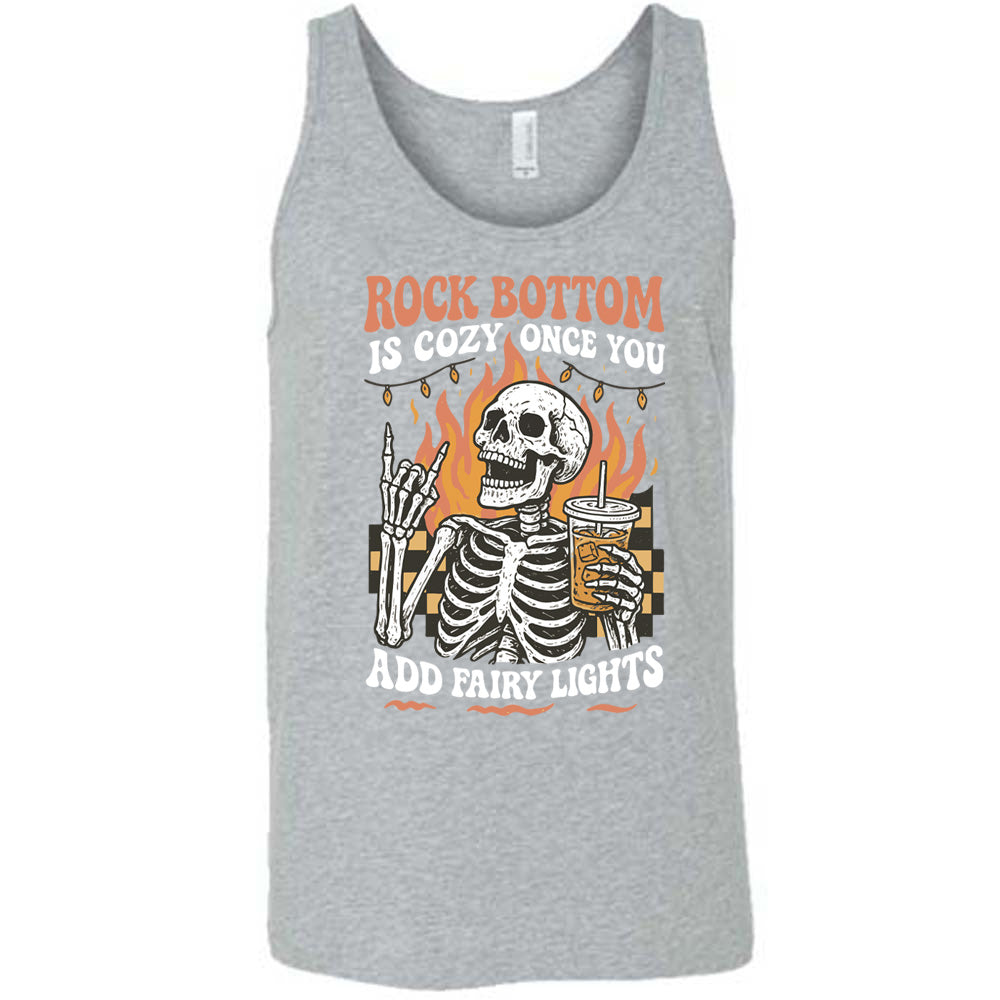 grey unisex tank with the words "rock bottom is cozy once you add fairy lights" with the graphic of a skeleton giving the rocker sign with one hand while holding an iced drink in the other, with a background of some checkerboard, some flames, and a string of fairy lights above the flames