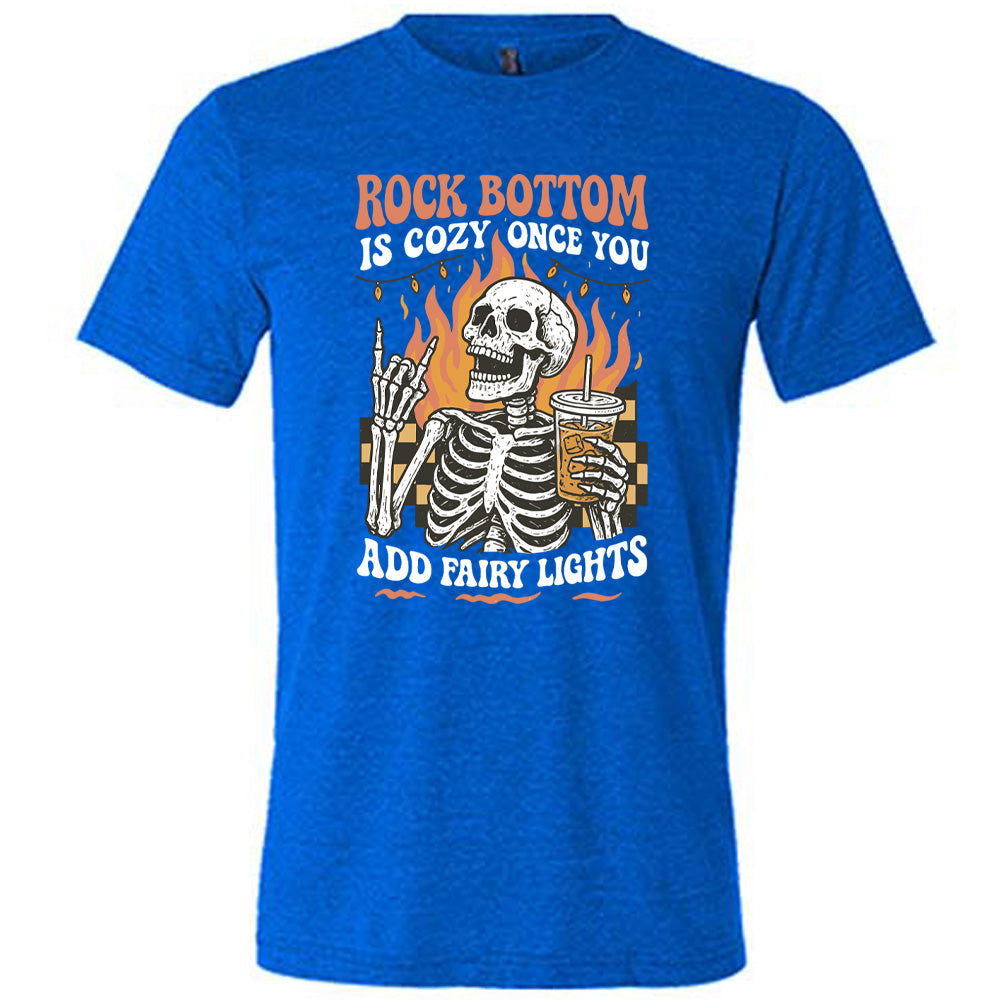 blue unisex tee with the words "rock bottom is cozy once you add fairy lights" with the graphic of a skeleton giving the rocker sign with one hand while holding an iced drink in the other, with a background of some checkerboard, some flames, and a string of fairy lights above the flames