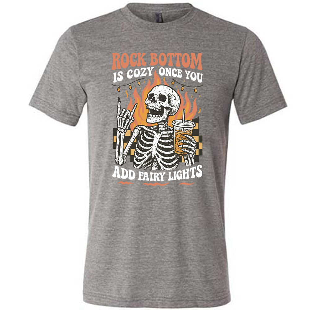 grey unisex tee with the words "rock bottom is cozy once you add fairy lights" with the graphic of a skeleton giving the rocker sign with one hand while holding an iced drink in the other, with a background of some checkerboard, some flames, and a string of fairy lights above the flames