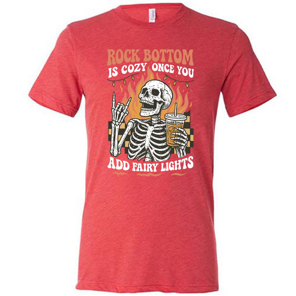 red unisex tee with the words "rock bottom is cozy once you add fairy lights" with the graphic of a skeleton giving the rocker sign with one hand while holding an iced drink in the other, with a background of some checkerboard, some flames, and a string of fairy lights above the flames