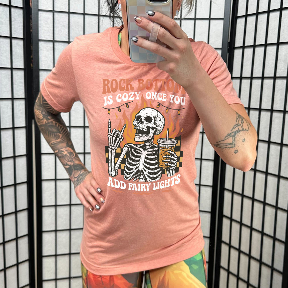 sunset unisex tee with the words "rock bottom is cozy once you add fairy lights" with the graphic of a skeleton giving the rocker sign with one hand while holding an iced drink in the other, with a background of some checkerboard, some flames, and a string of fairy lights above the flames