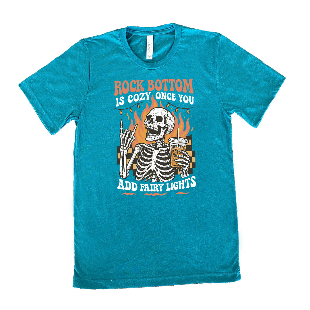 teal unisex tee with the words "rock bottom is cozy once you add fairy lights" with the graphic of a skeleton giving the rocker sign with one hand while holding an iced drink in the other, with a background of some checkerboard, some flames, and a string of fairy lights above the flames