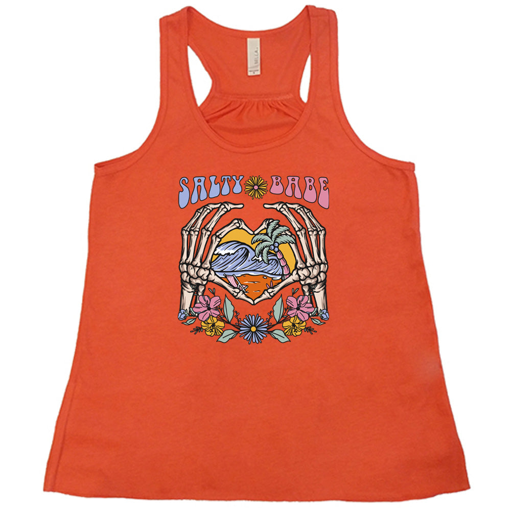 Coral colored flowy tank with a skeleton heart hands around ocean waves graphic on it that says "Salty Babe" 