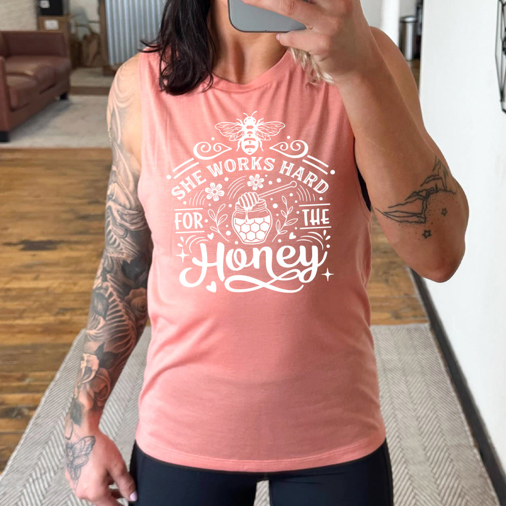 peach muscle tank with the words "she works for the honey" surrounded by accenting curved lines, with flowers, dots, stars, and hears scattered around, and a honeypot and spoon in the center, and a bee on the top