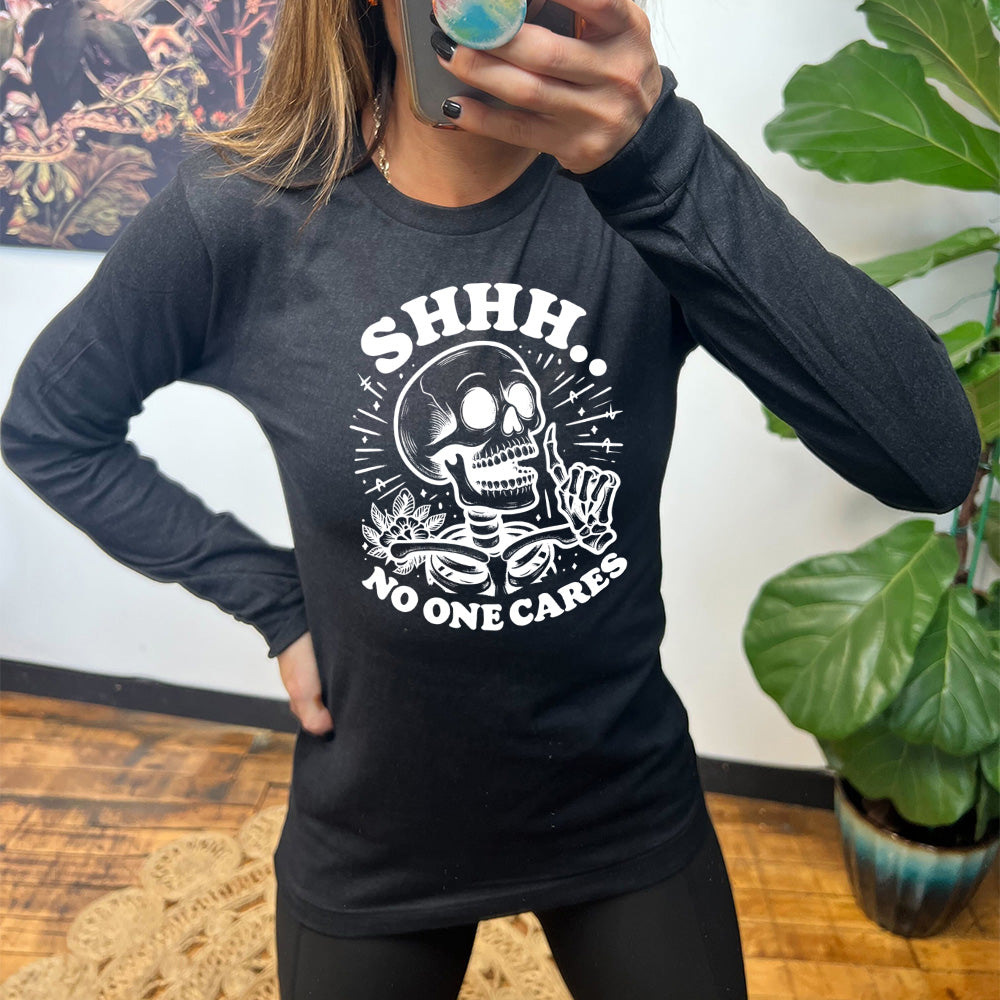 heather black long sleeve with the words "Shhh.. no one cares" on it, with the graphic of a skeleton holding a finger to its lips 
