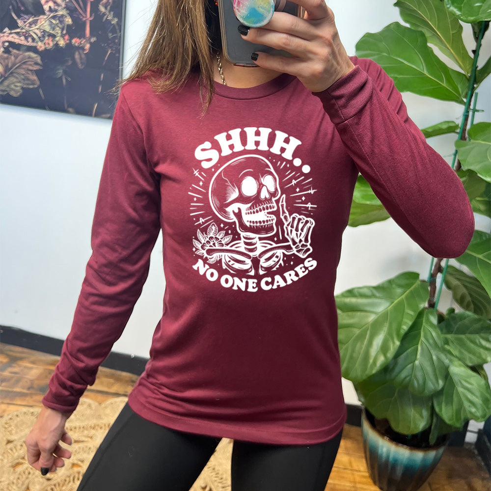 heather cardinal long sleeve with the words "Shhh.. no one cares" on it, with the graphic of a skeleton holding a finger to its lips 