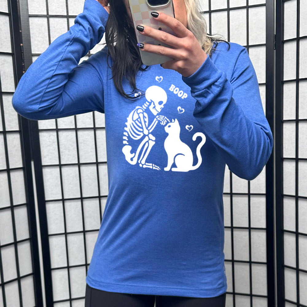 heather true royal long sleeve shirt with the graphic of a sitting skeleton touching the head of a sitting cat, with the word "boop" above this action, and four little hearts around them