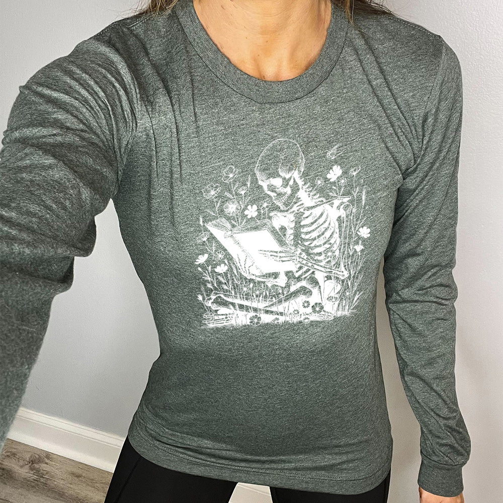 heather forest long sleeve shirt with the graphic of a skeleton reading a book, sitting cross-legged in a bed of tall flowers