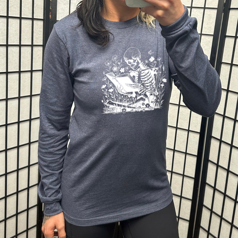 heather navy long sleeve shirt with the graphic of a skeleton reading a book, sitting cross-legged in a bed of tall flowers