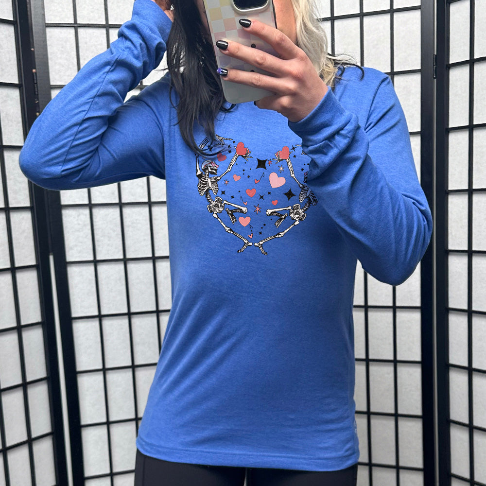 heather true royal long sleeve shirt with the graphic of two skeletons creating the shape of a heart with their bodies, appearing to be dancing, with many hearts between them, and a few little stars