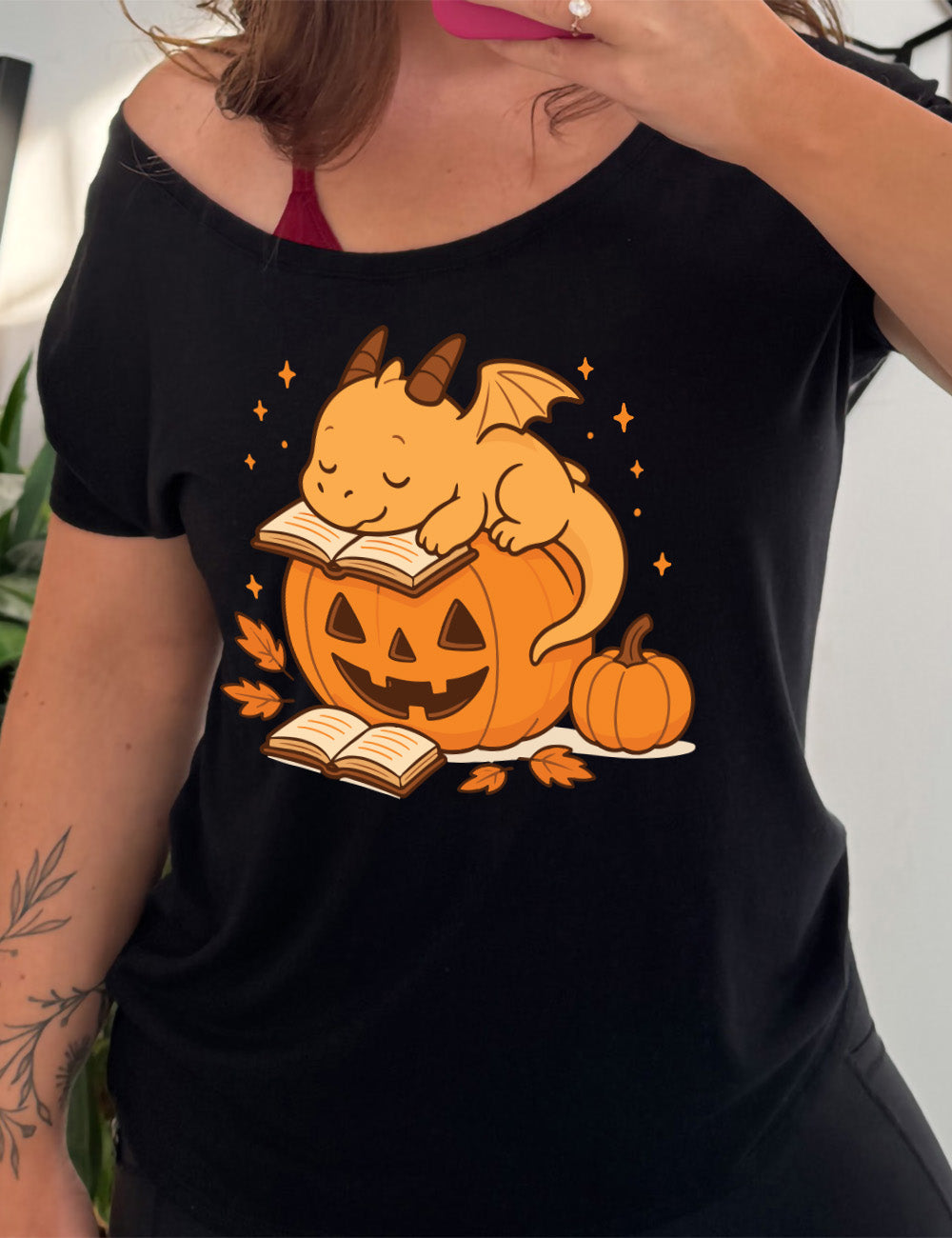 black slouchy tee with the graphic of a cartoon-style dragon sleeping, with its head in its open book, on top of a jack-o-lantern, with another open book and small pumpkin next to all this, and a scattering of stars, and a few leaves
