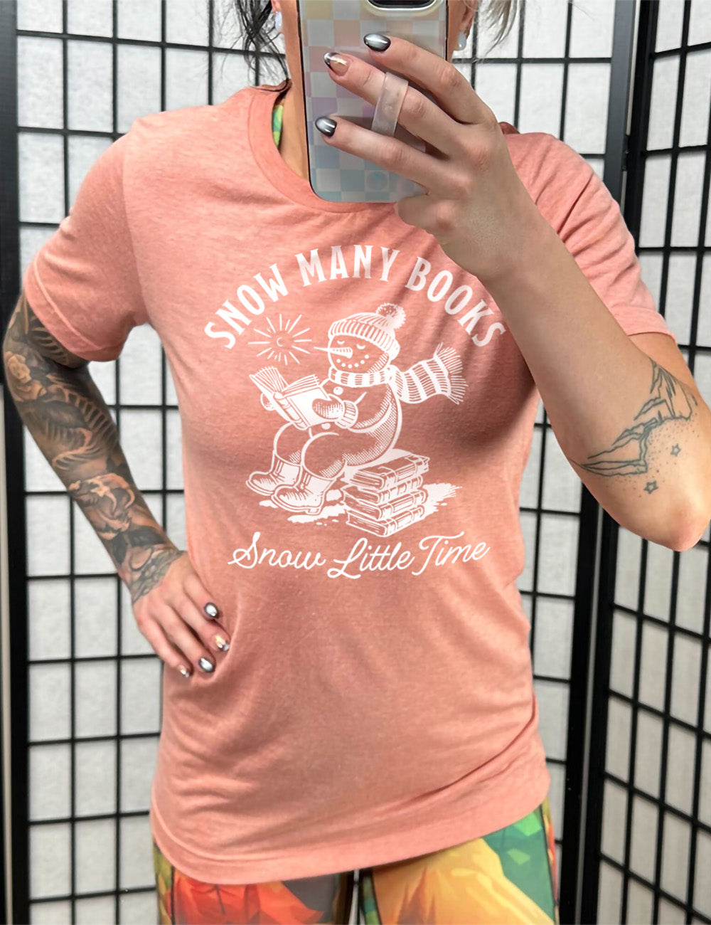 sunset unisex tee with the graphic of a happy snowman, wearing a snowsuit and boots, mittens, a hat, and  book, reading a book while sitting on a stack of books, with a sun in the sky, and the words arching above saying "snow many books" and the words in a line of cursive below saying "snow little time"