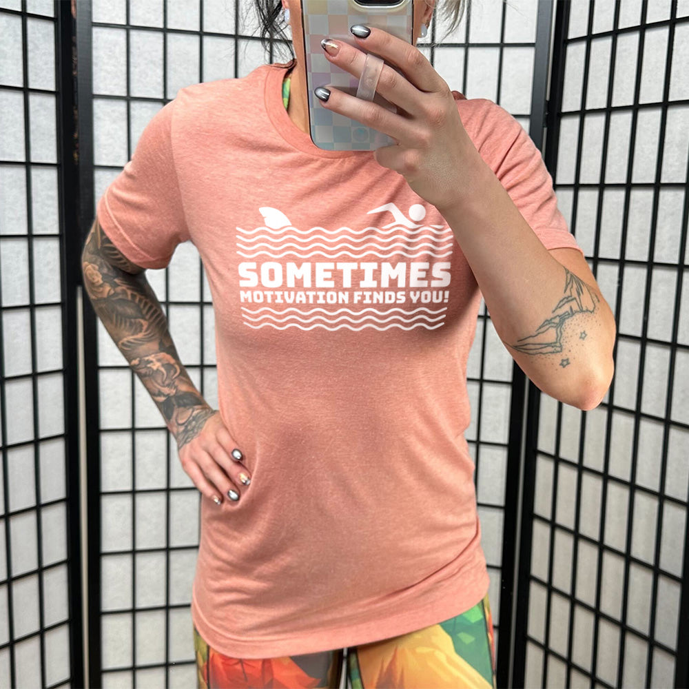 sunset unisex tee with the words "sometimes motivation finds you!" in between the lined graphic of waves of the sea, and on top of the waves is the graphic of a figure swimming away from a fin in the water behind