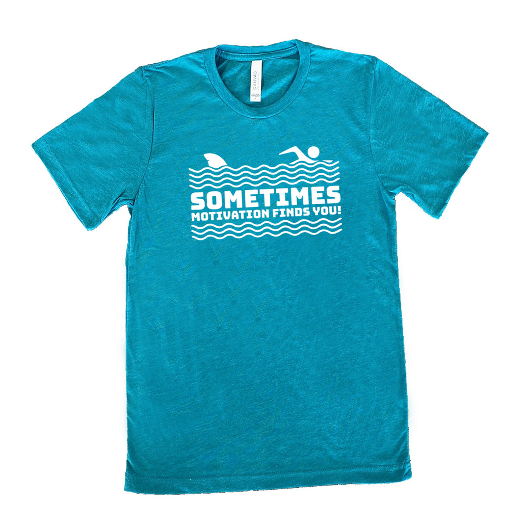 teal unisex tee with the words "sometimes motivation finds you!" in between the lined graphic of waves of the sea, and on top of the waves is the graphic of a figure swimming away from a fin in the water behind
