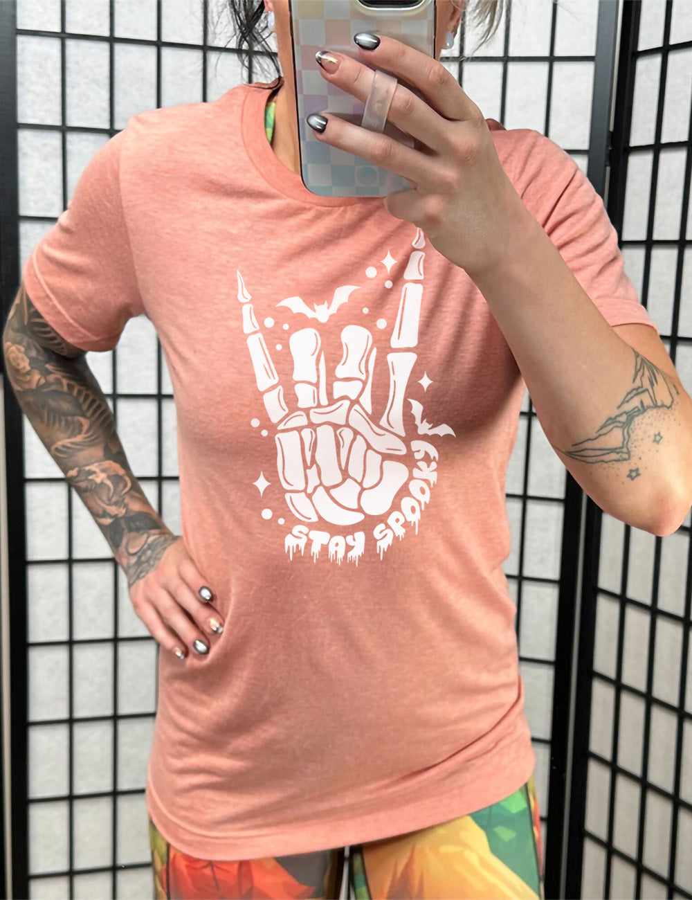 sunset unisex tee with the large graphic of a rocker skeletal hand, with two little bats, and few stars and dots, with the words underneath saying "stay spooky"