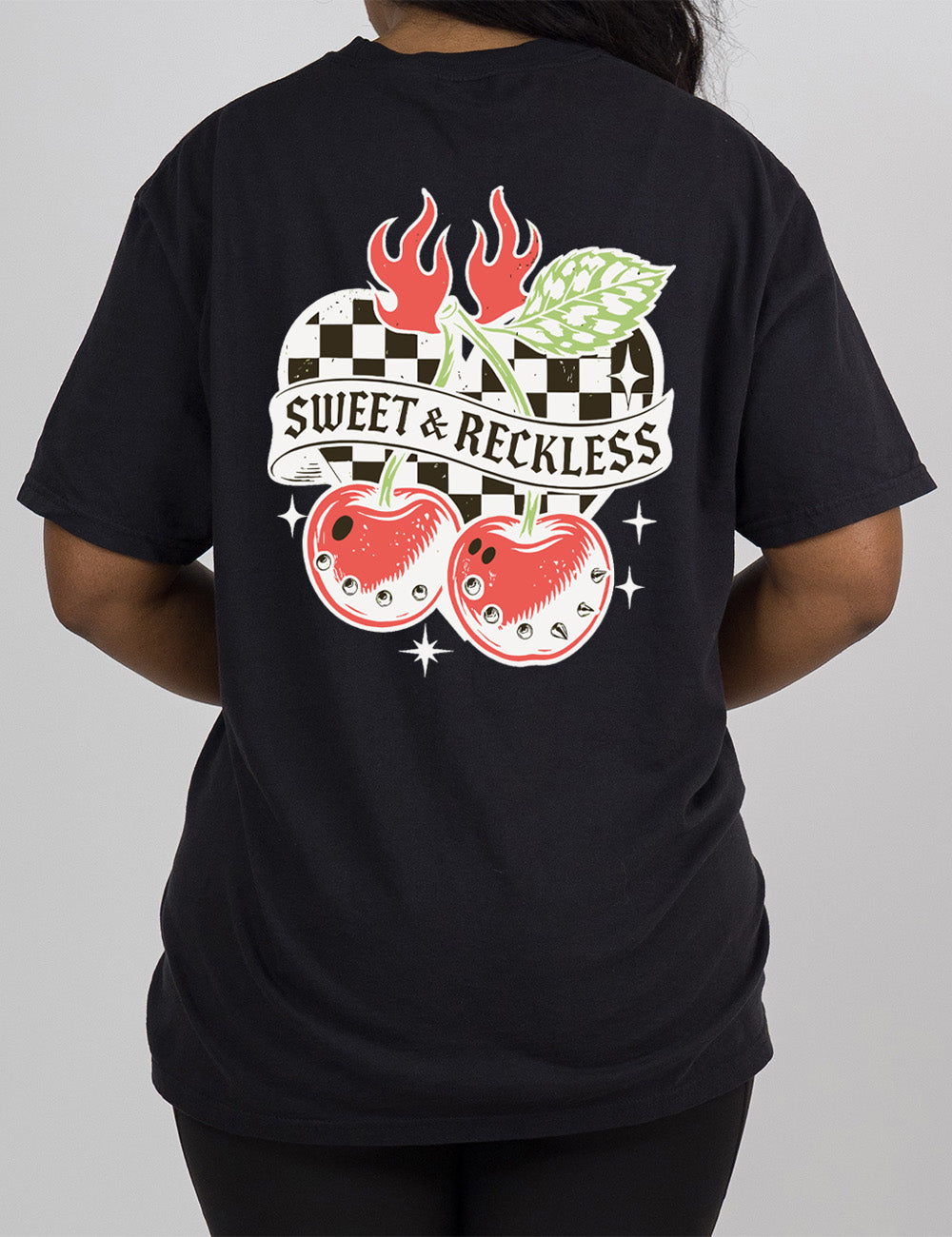 black modern tee with print placement on the back, with the graphic of two large cherries, with spikes, on connected stems with a leaf, in front of a checkered heart, with flames on top, a few stars around, with a banner across the heart saying "sweet & reckless"
