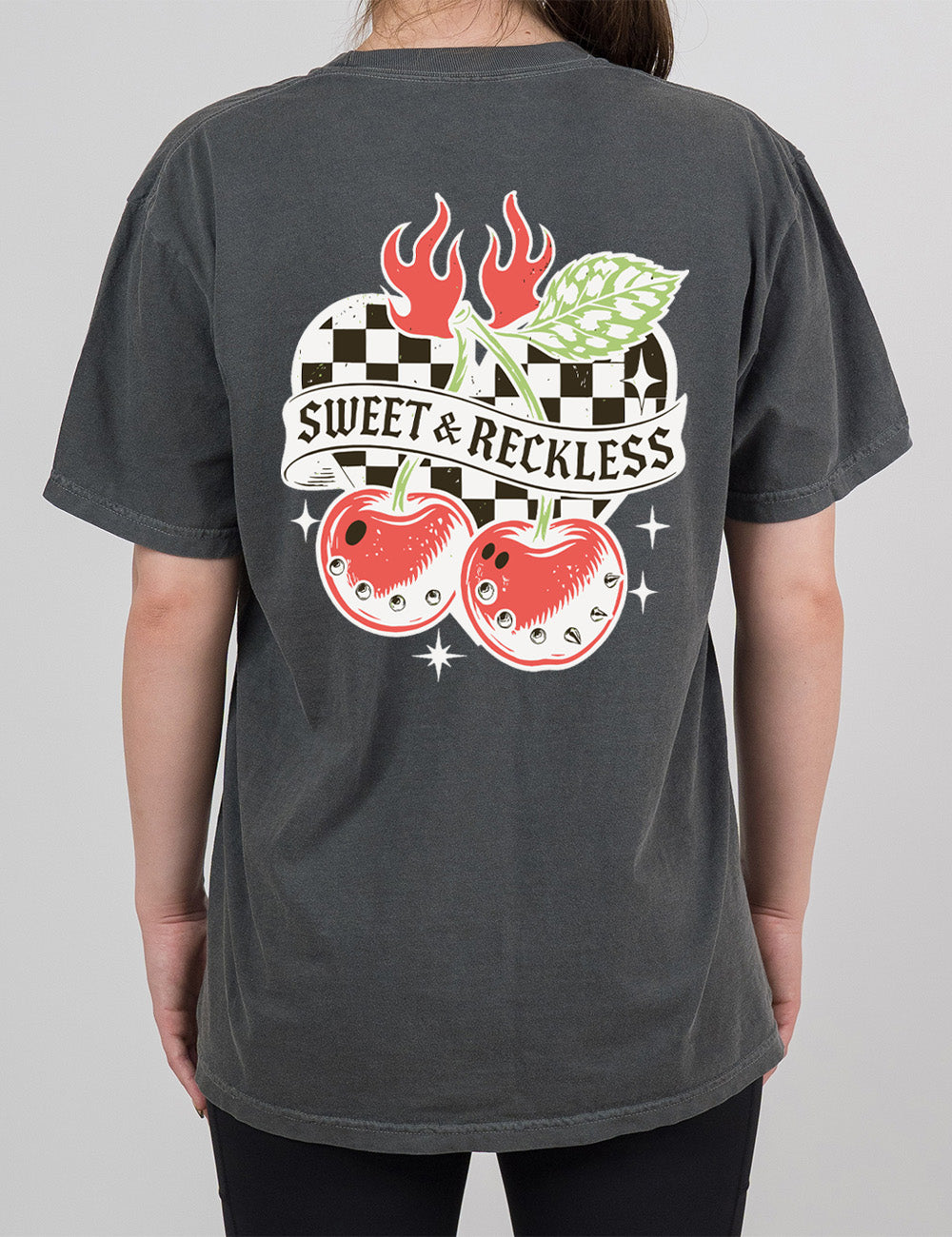 pepper modern tee with print placement on the back, with the graphic of two large cherries, with spikes, on connected stems with a leaf, in front of a checkered heart, with flames on top, a few stars around, with a banner across the heart saying "sweet & reckless"