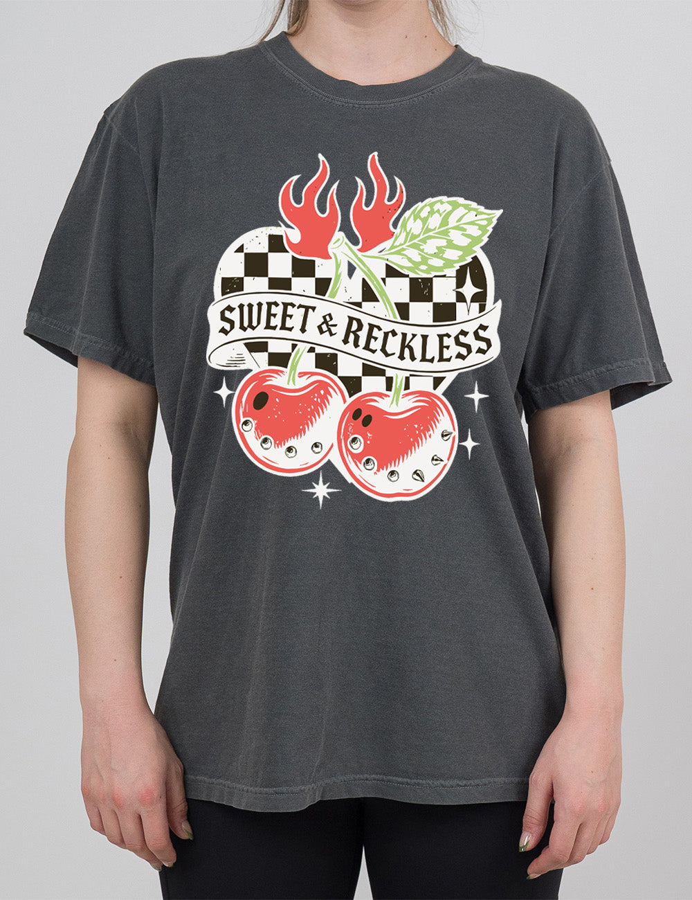 pepper modern tee with print placement on the front, with the graphic of two large cherries, with spikes, on connected stems with a leaf, in front of a checkered heart, with flames on top, a few stars around, with a banner across the heart saying "sweet & reckless"