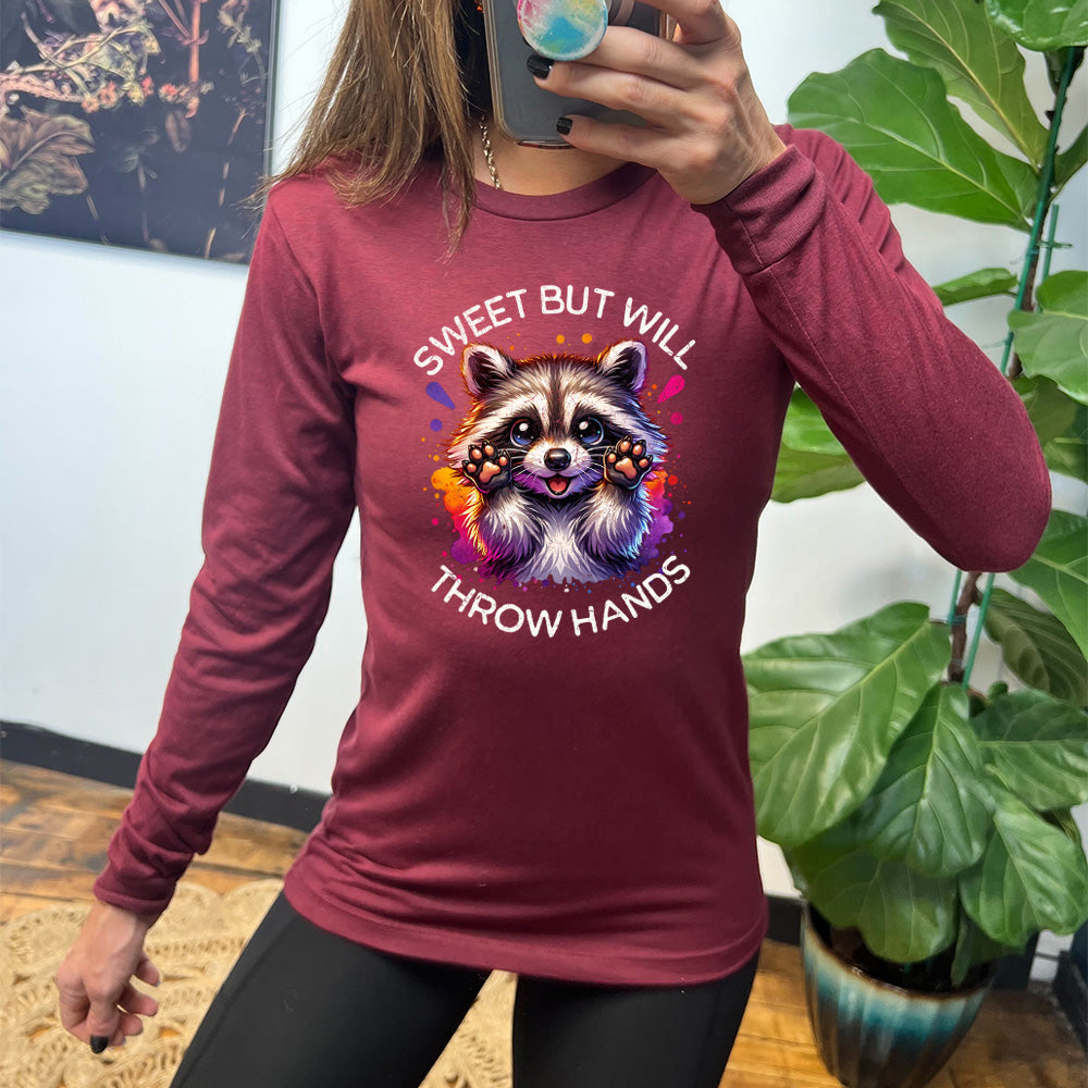 heather cardinal long sleeve shirt with the words "sweet but will throw hands" on it, with the graphic cartoon of a racoon with innocent-looking eyes, holding up its paws