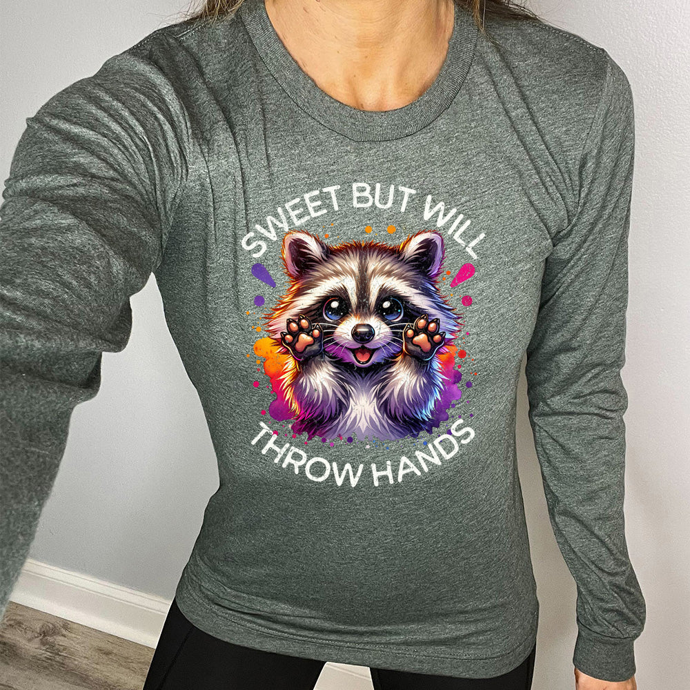 heather forest long sleeve shirt with the words "sweet but will throw hands" on it, with the graphic cartoon of a racoon with innocent-looking eyes, holding up its paws