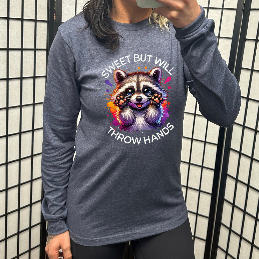 heather navy long sleeve shirt with the words "sweet but will throw hands" on it, with the graphic cartoon of a racoon with innocent-looking eyes, holding up its paws