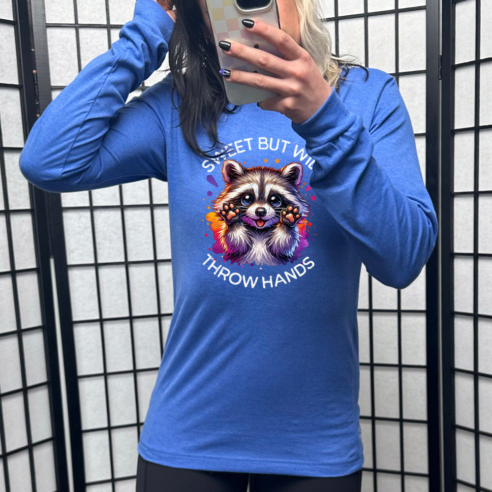 heather true royal long sleeve shirt with the words "sweet but will throw hands" on it, with the graphic cartoon of a racoon with innocent-looking eyes, holding up its paws