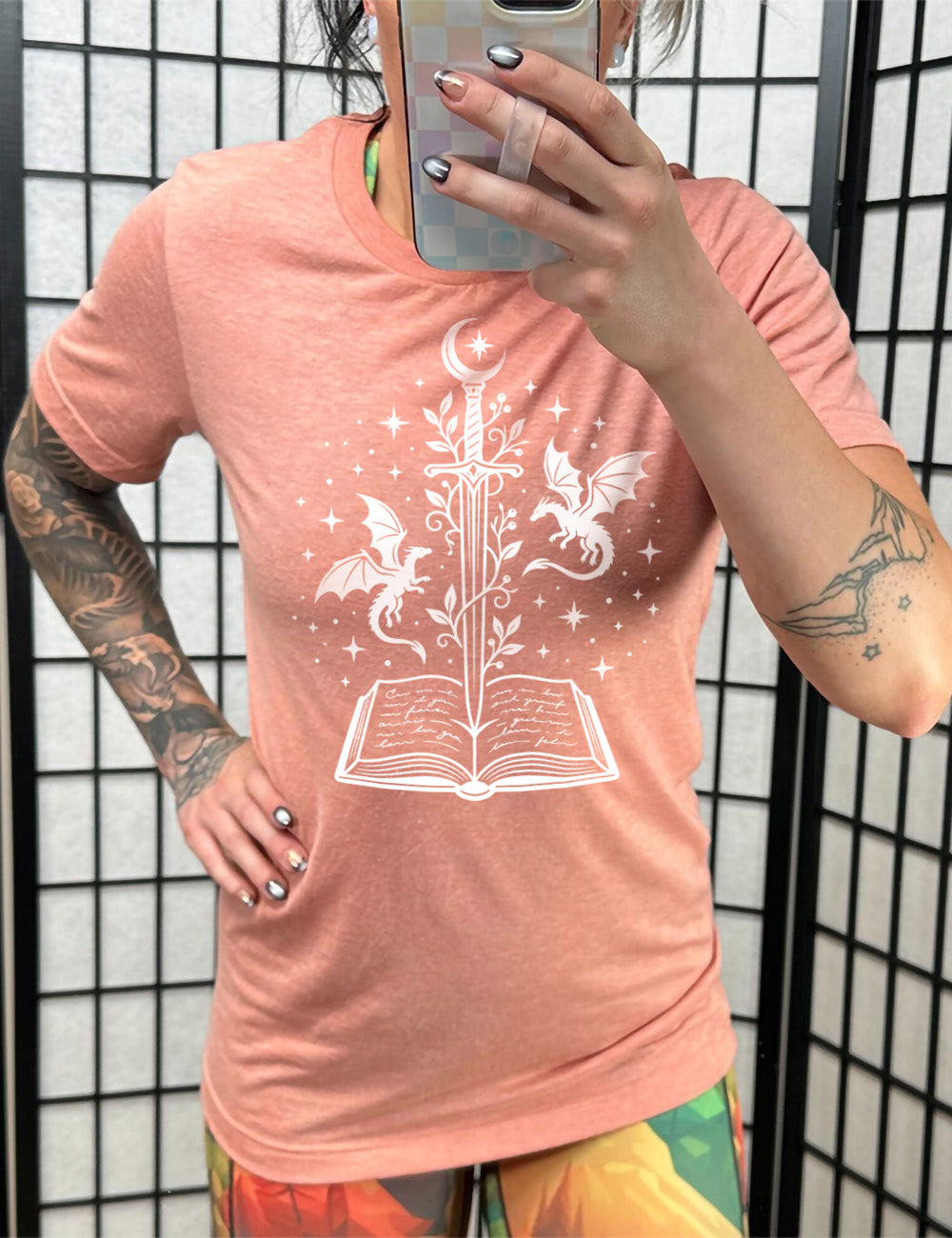 sunset unisex tee with the graphic of a sword, with a crescent moon on top of the handle and with a vine wrapping around it, pointing down into an open book, surrounded by stars, and a dragon flying on either side