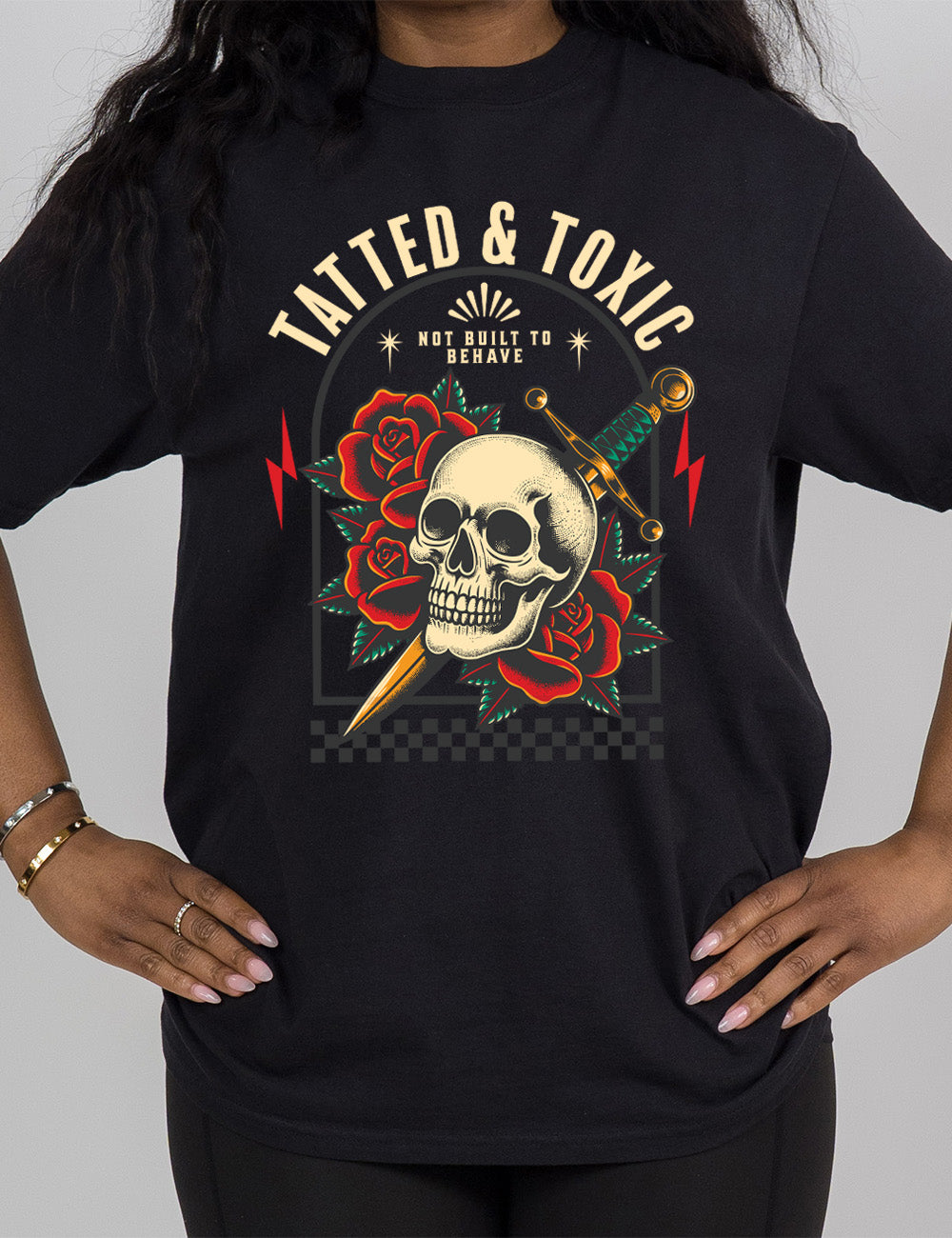 black modern tee with the print placement being on the front, with the words "tatted & toxic" on it, with the graphic of a skull, with a knife through its head, among roses, with small words above it, saying "not built to behave," within an arched box, over a strip of checkered design, with a lightning bolt on either side