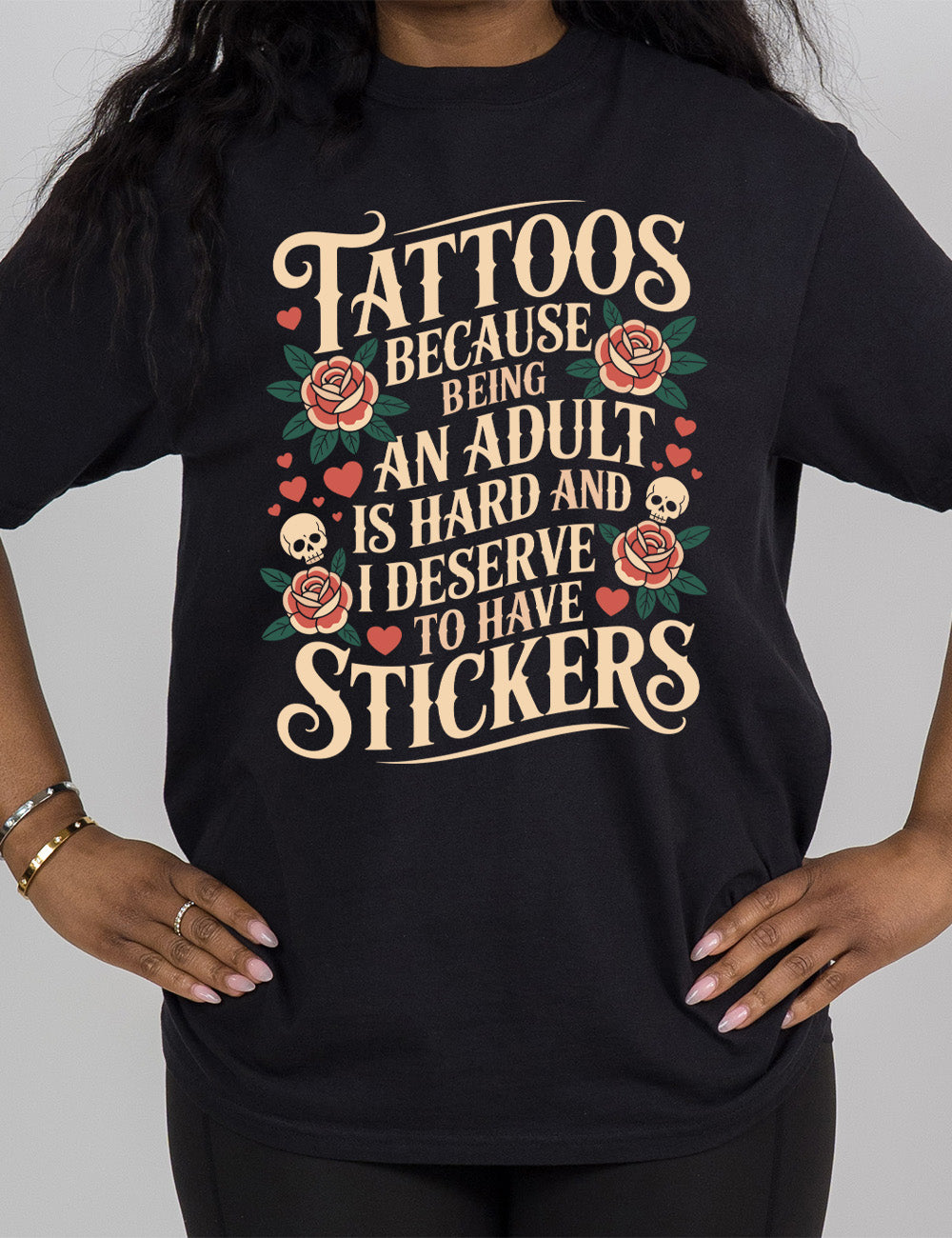 black modern tee with print placement on the front, with the words "tattoos because being an adult is hard and I deserve to have stickers" on it, with the graphics of roses, hearts, and skulls on the side of the words
