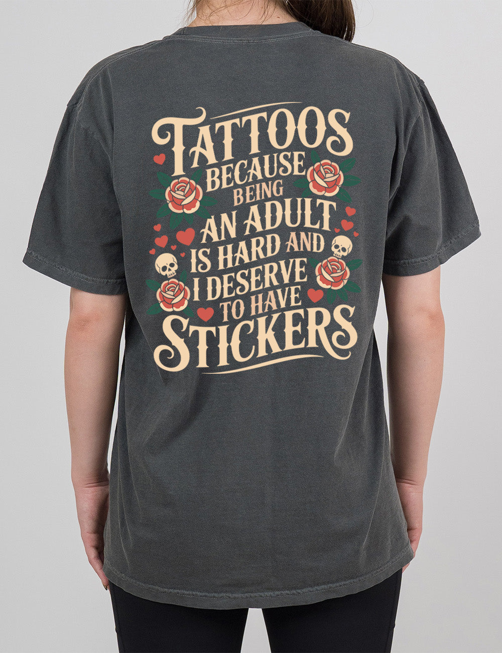 pepper modern tee with print placement on the back, with the words "tattoos because being an adult is hard and I deserve to have stickers" on it, with the graphics of roses, hearts, and skulls on the side of the words