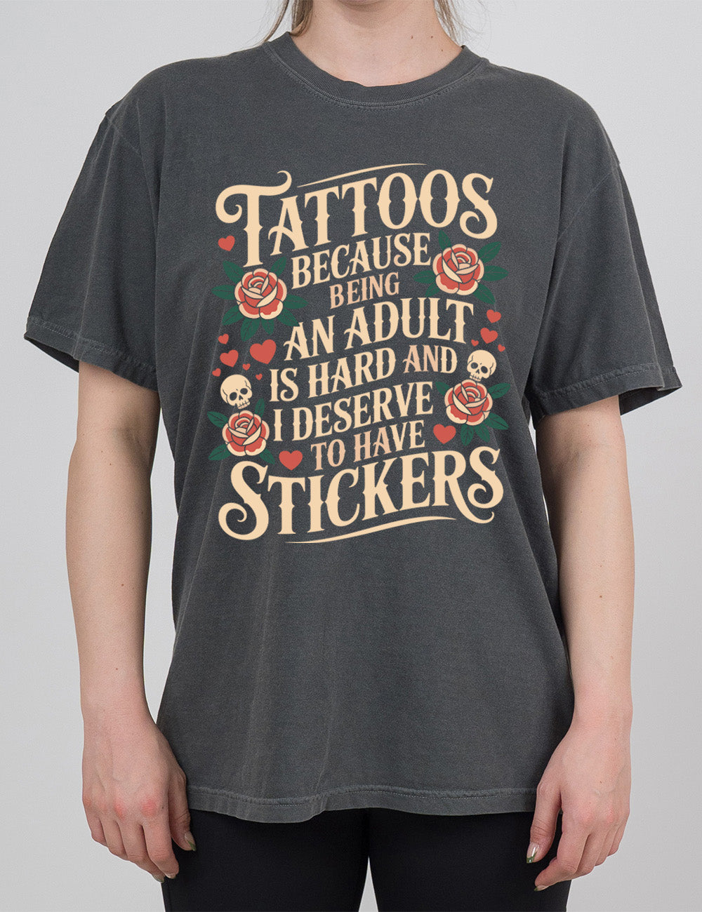 pepper modern tee with print placement on the front, with the words "tattoos because being an adult is hard and I deserve to have stickers" on it, with the graphics of roses, hearts, and skulls on the side of the words