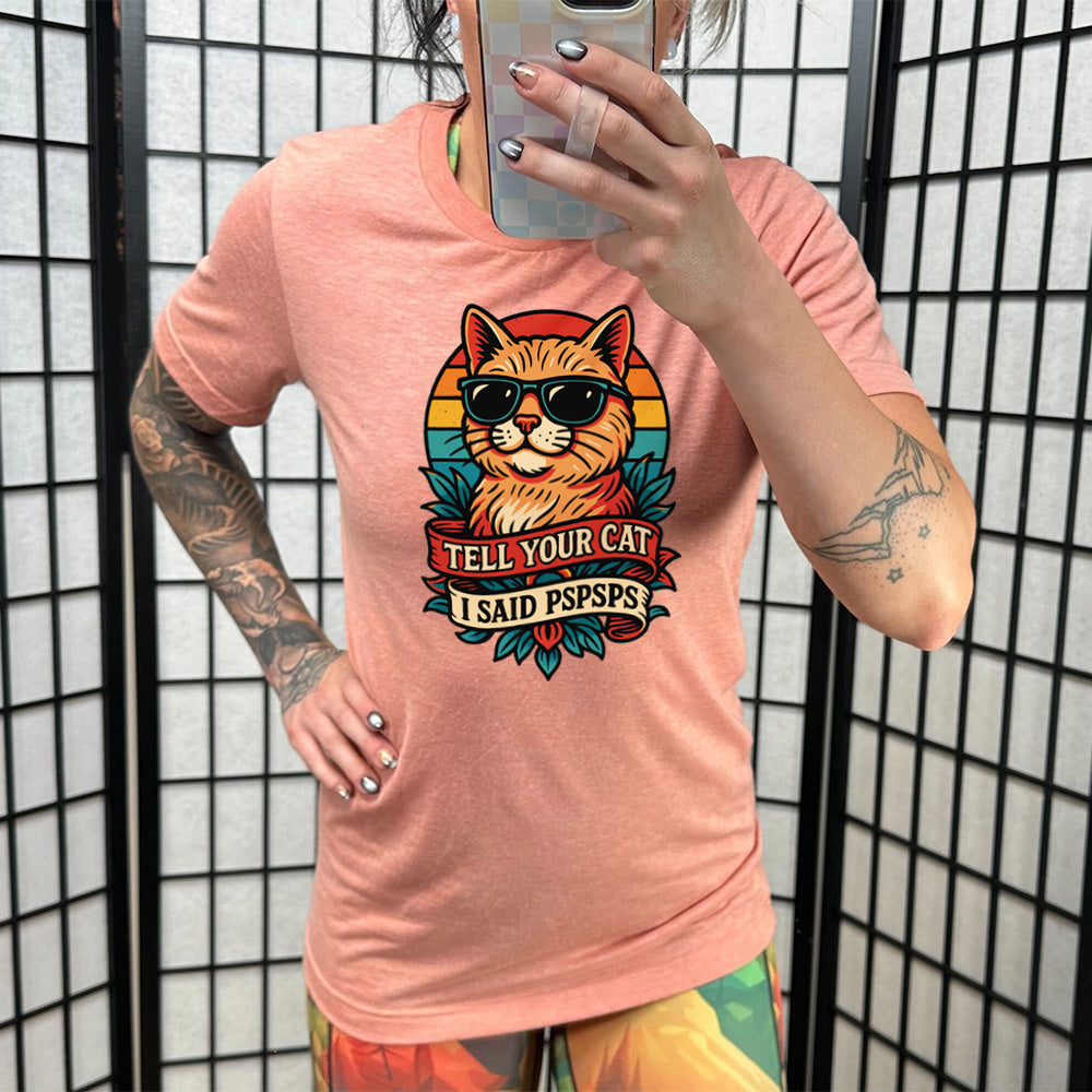 sunset unisex tee with the large graphic of a cat head and torso, wearing sunglasses, with a colorful background, and two banners below it with the words "tell your cat I said pspsps" on them