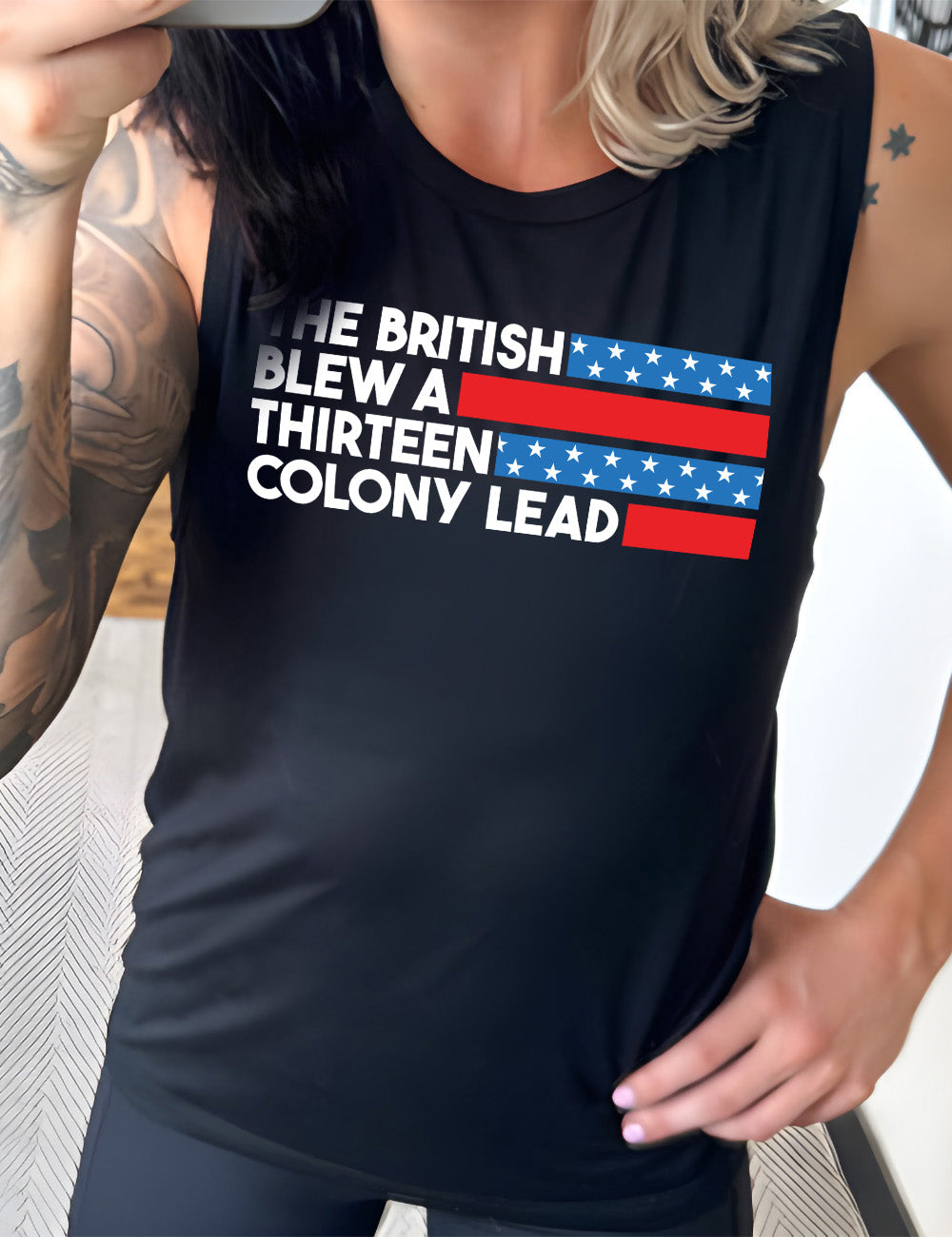 black muscle tank with the words "the British" (and a blue starred bar finishing the space to the right), then below "blew a" (and a red bar), then below "thirteen" (and a blue starred bar),  and below "colony lead" (and a red bar)